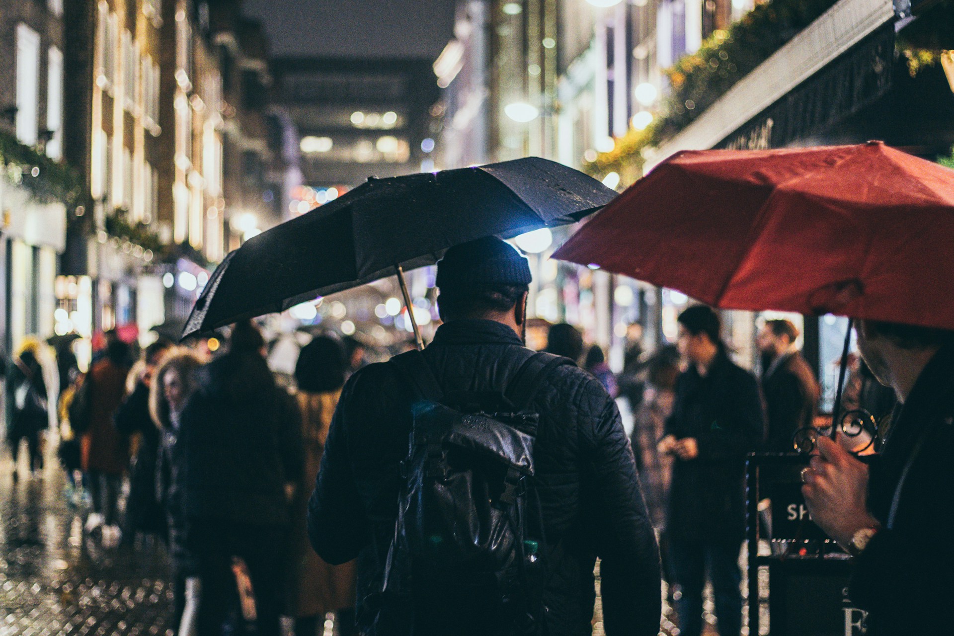 Man carrying a black EDC umbrella through a busy, rain-soaked city street at night.