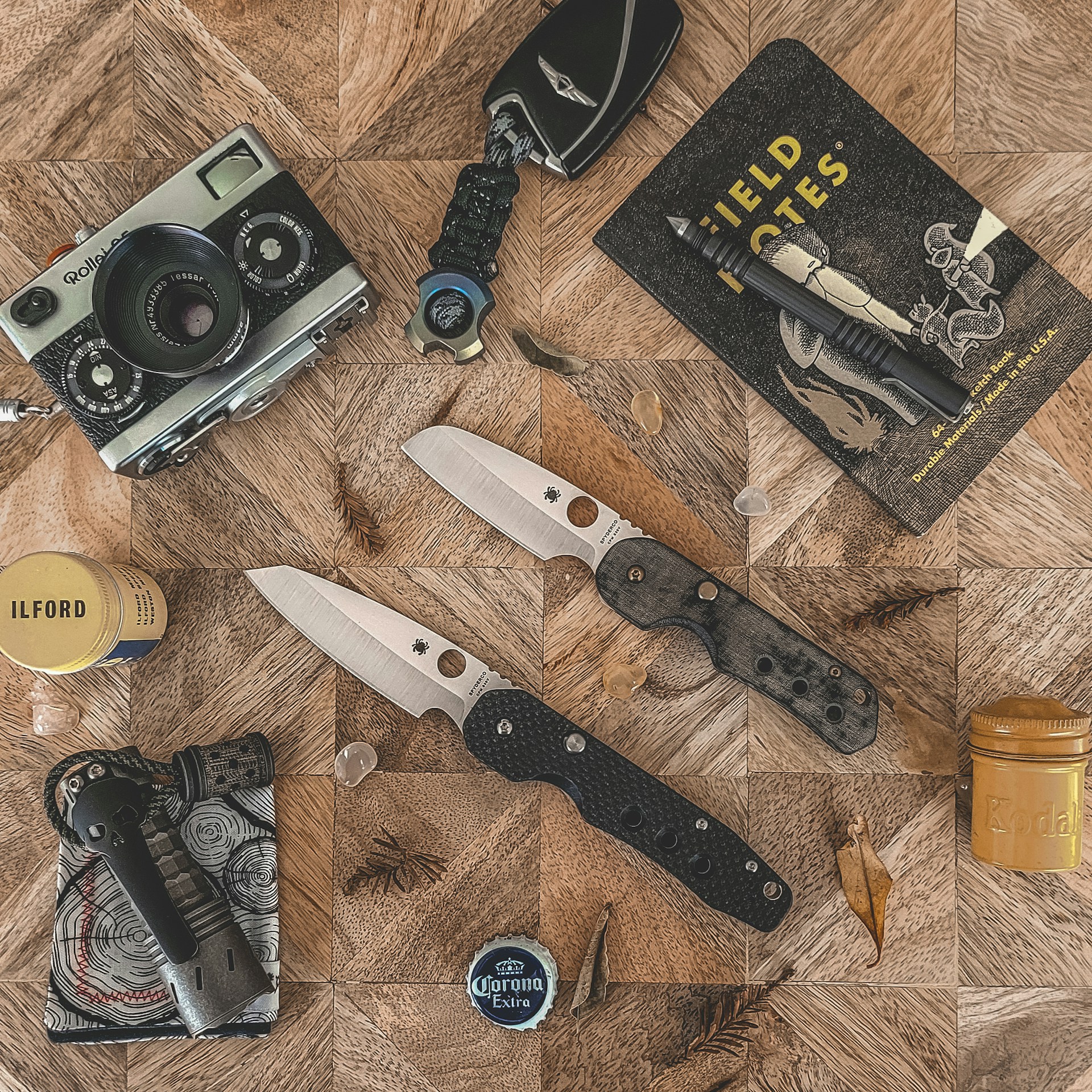 A flat lay of everyday carry (EDC) gear including two Spyderco folding knives, a vintage Rollei camera, a tactical pen, a Field Notes notebook, a keychain tool, a flashlight, and film canisters, arranged on a wooden surface.